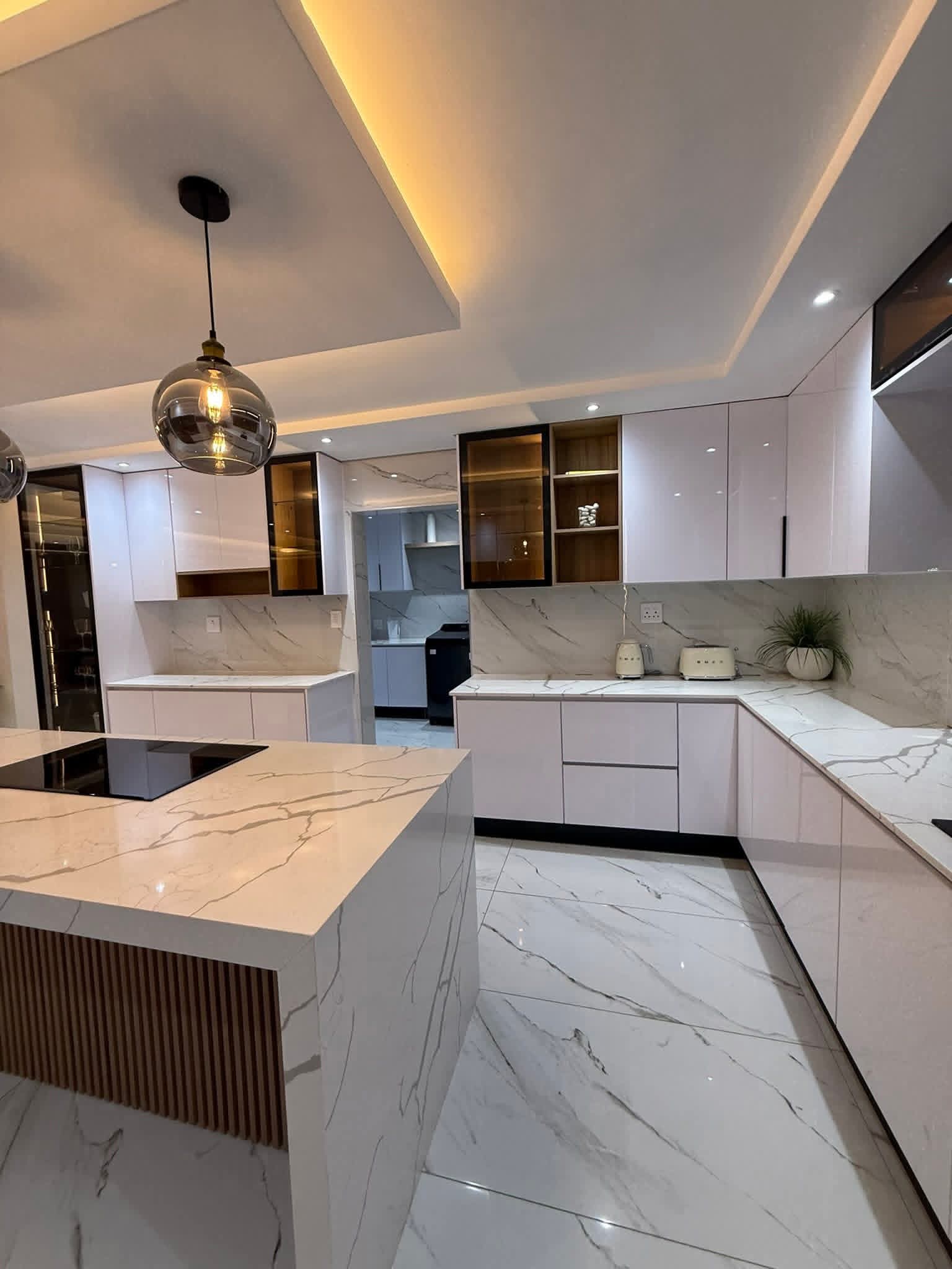 White kitchen with marble worktops, smoked-glass pendants and warm cove lighting in a tray ceiling