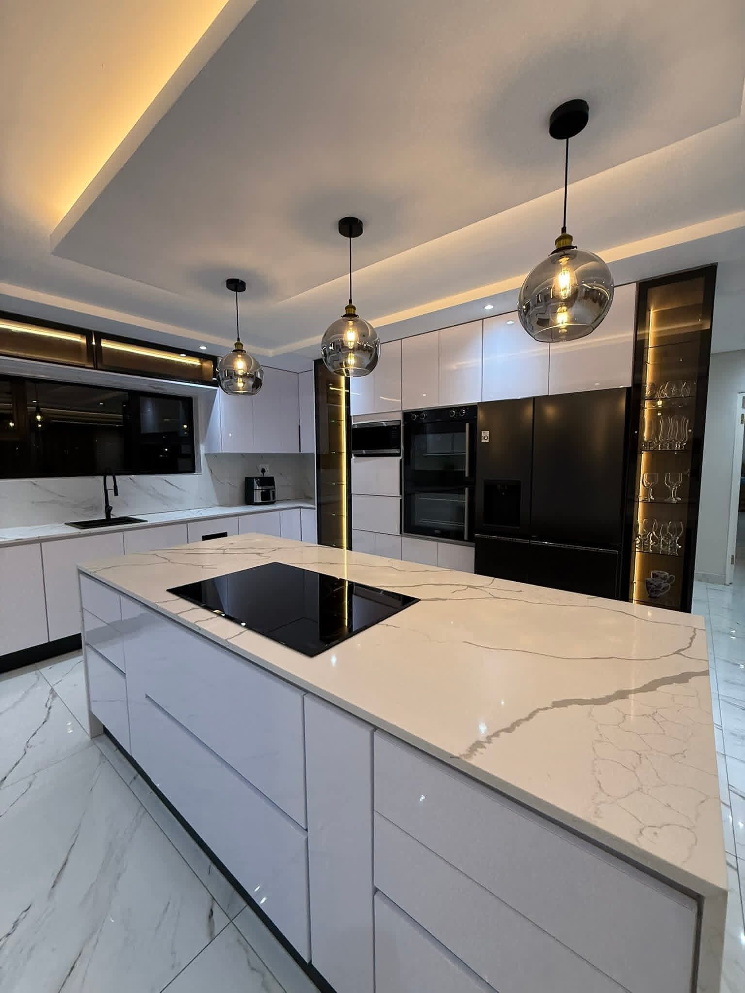 Evening kitchen with marble island, globe pendants, black appliances and lit glass display cabinets